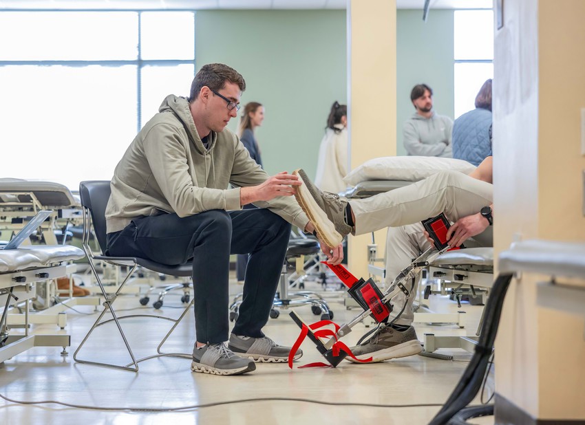 Student practices a lower-leg orthopedic assessment on a classmate in a physical therapy lab, using measurement equipment around the ankle and foot.