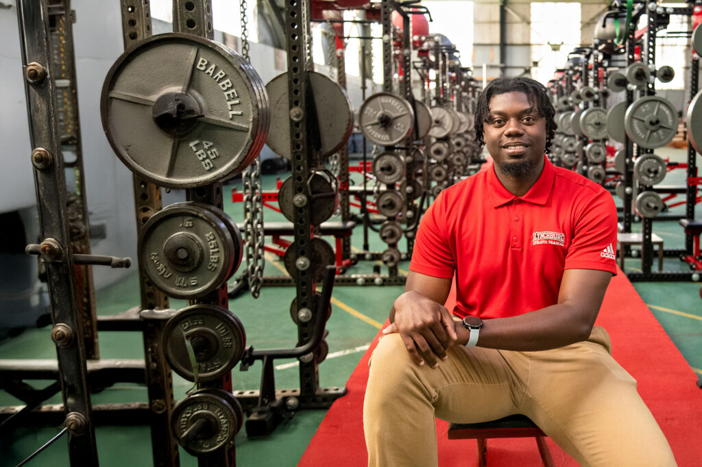 Top Athletic Training Grad Programs: University of Lynchburg athletic training staff member seated in a strength training facility with weight racks and plates in the background.