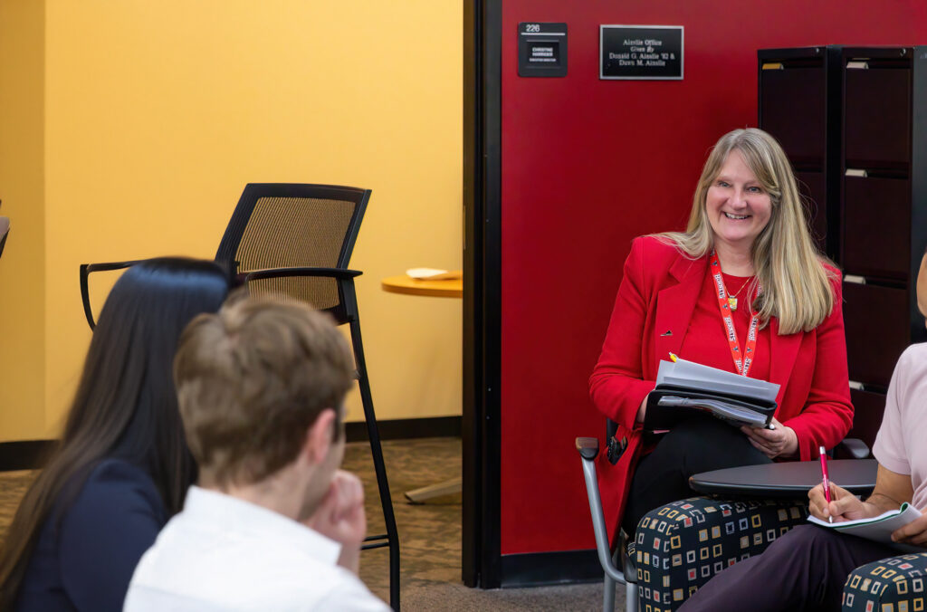 Staff member in a red blazer meets with students in a small group setting, holding papers and smiling during discussion.