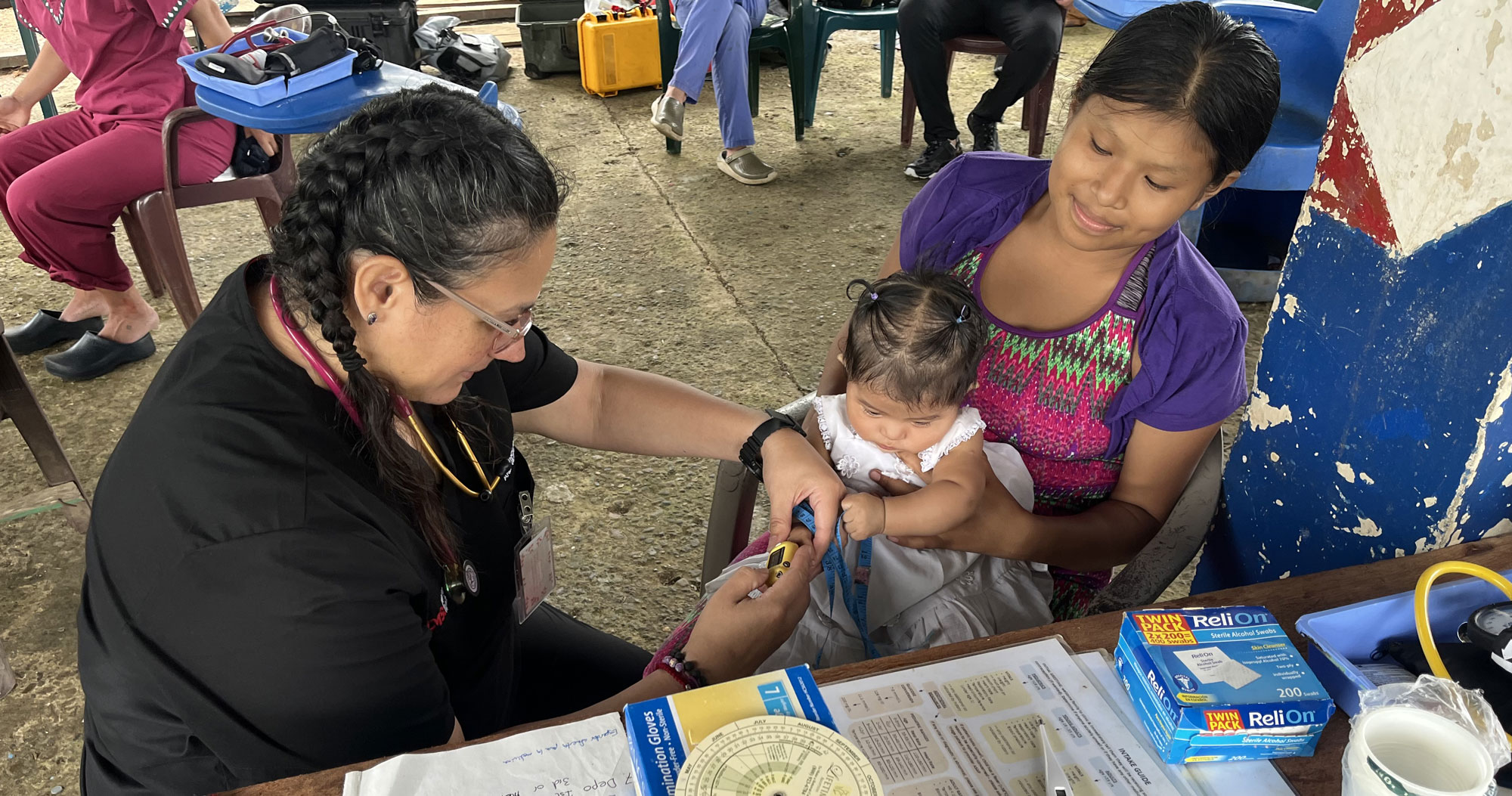 Healthcare professional uses a measuring tape to assess an infant while the child’s mother holds the baby during a community medical mission clinic.