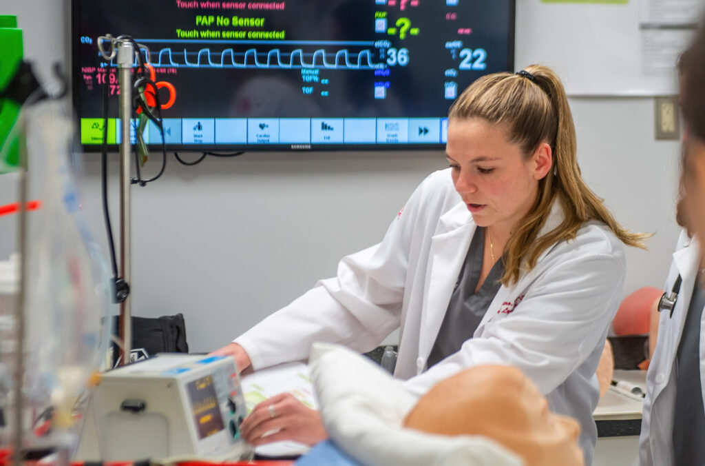 Physician assistant studies student in a white coat reviews patient monitoring data during a clinical skills lab exercise.