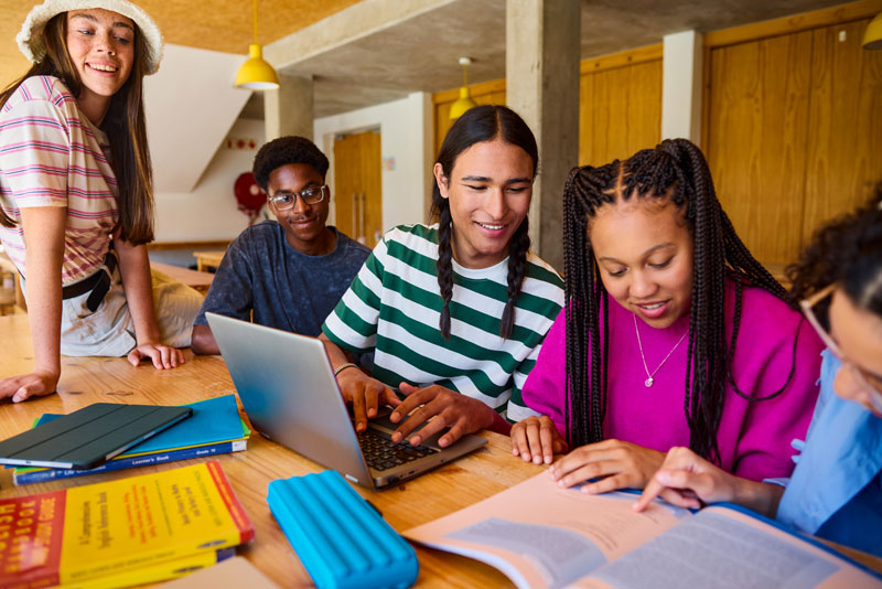 Group of college students study together at a table, with one student using a laptop while others read and discuss notes.