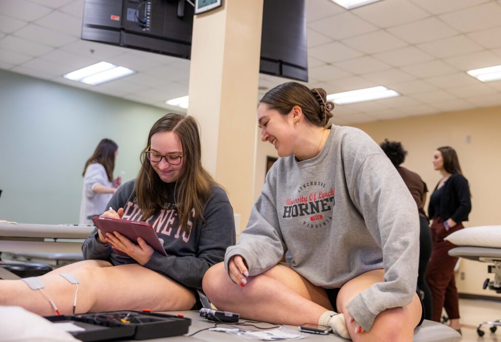 Two students work together during a physical therapy lab exercise, recording data while electrodes and testing equipment are attached to a classmate’s leg.